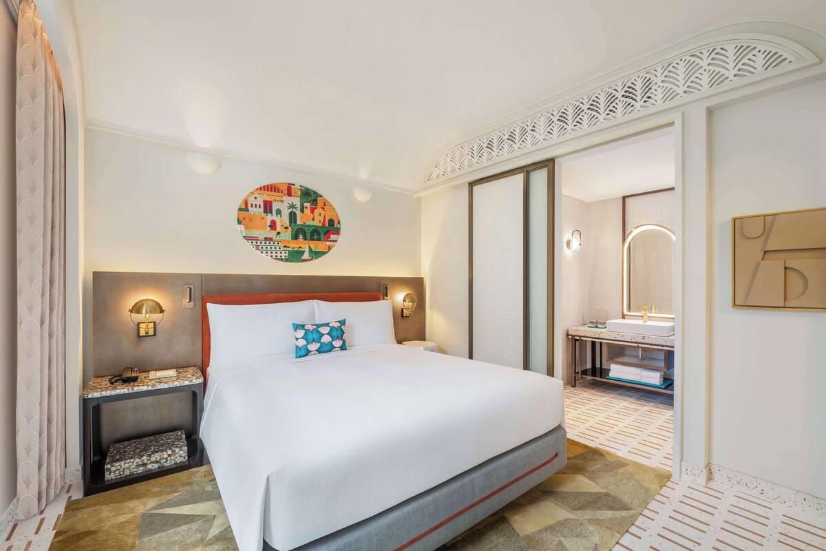 A hotel room with a large bed, a nightstand with a lamp, a bathroom with a mirror and sink, and a decorative wall piece.
