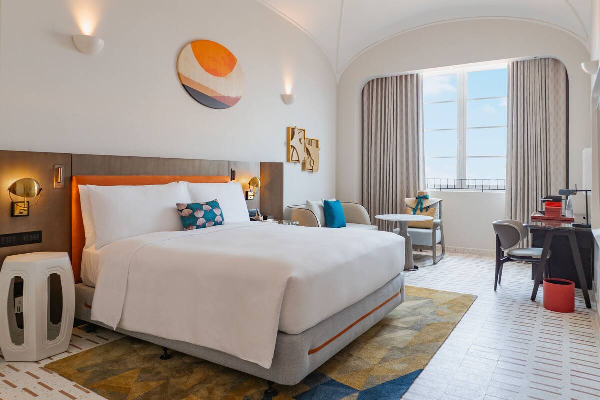 A hotel room with a large bed, a desk, a chair, and a window with curtains.