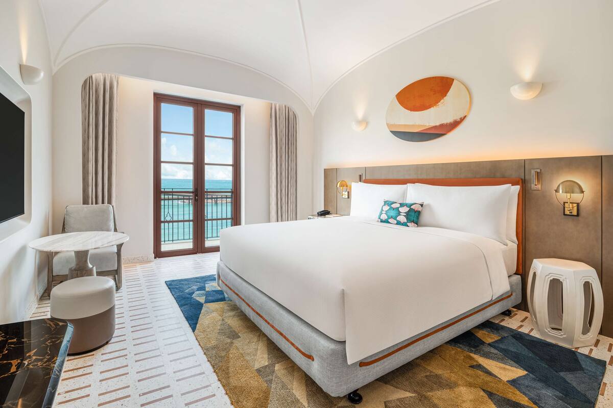 A hotel room with a large bed, a small round table, a chair, a sofa, a cat litter box, and a view of the ocean through a sliding door.