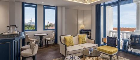 Habitación de hotel moderna con sofá, sillas, zona de bar y ventanas amplias con vistas a la ciudad.