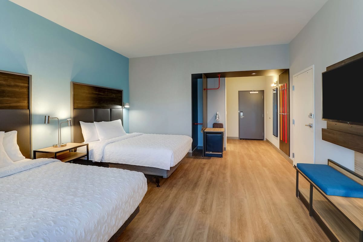 A hotel room with two beds, a TV, a bench, and a bathroom visible through an open door.