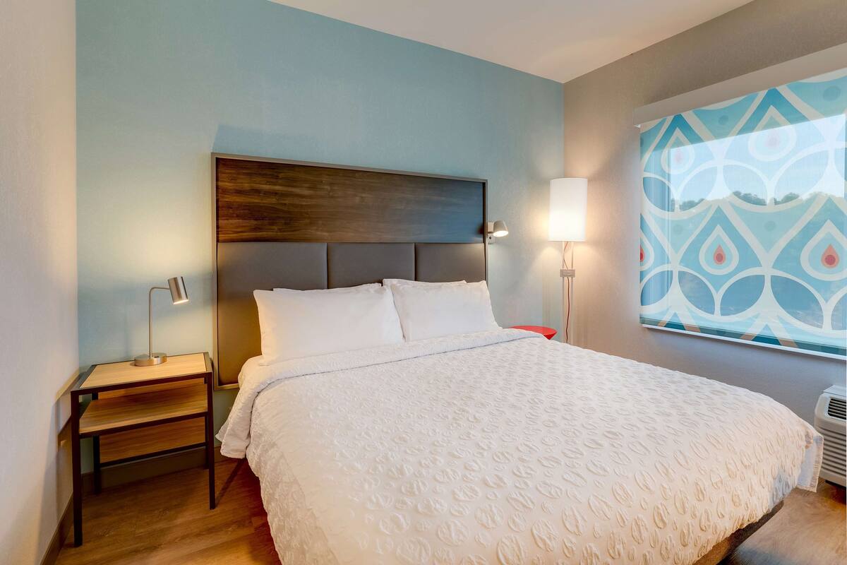A hotel room with a large bed, a nightstand with a lamp, and a wall-mounted air conditioner.