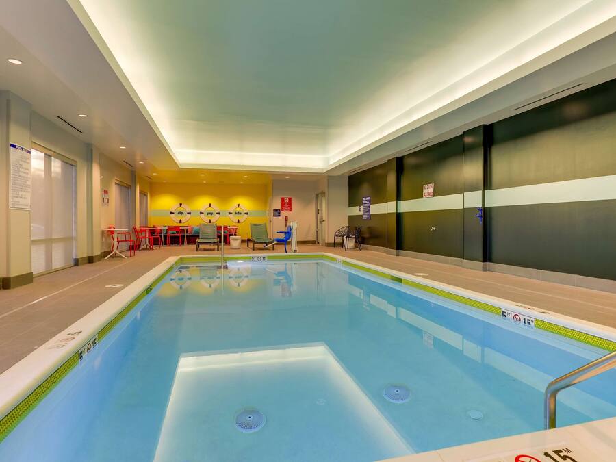 Una piscina interior con superficie de agua cristalina, rodeada por un suelo y paredes de azulejos. Hay sillas y mesas con una pared amarilla de fondo.