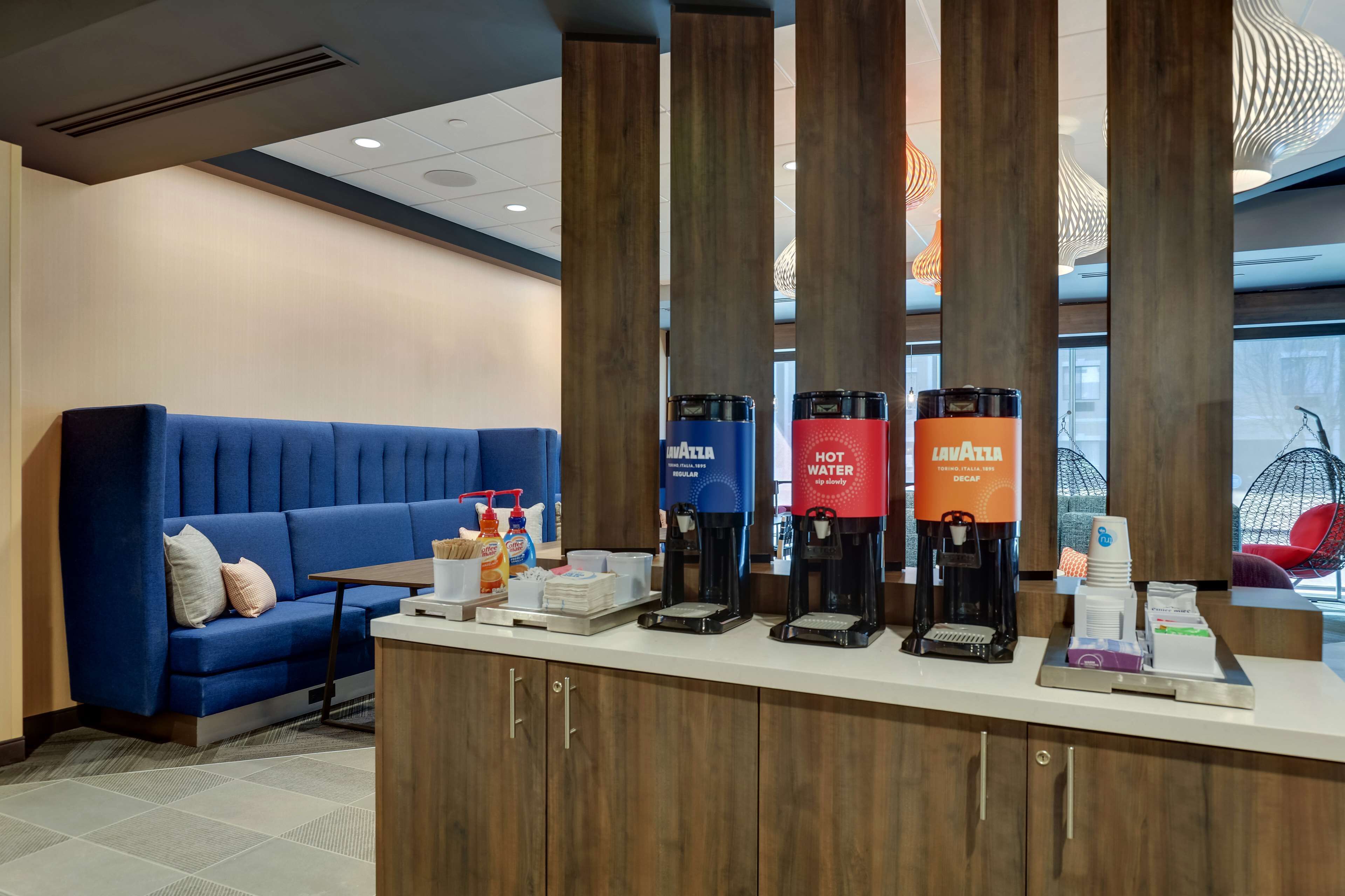 Zona de recepción de hotel con un sofá azul, un rincón de café con dispensadores de marca y una barra con diversos servicios.