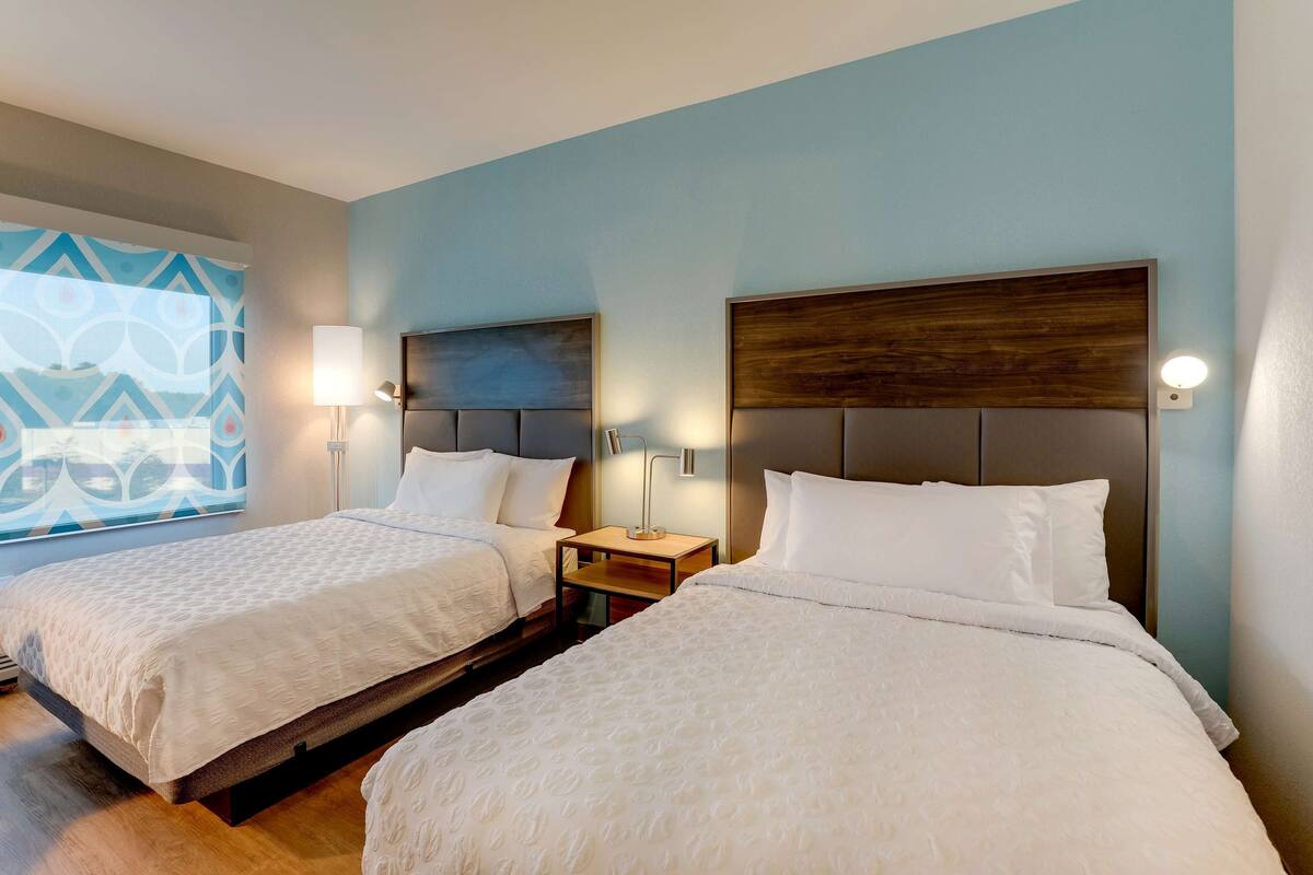 A hotel room with two beds, each with a wooden headboard and white bedding. There are bedside tables with lamps and a window with a geometric pattern.