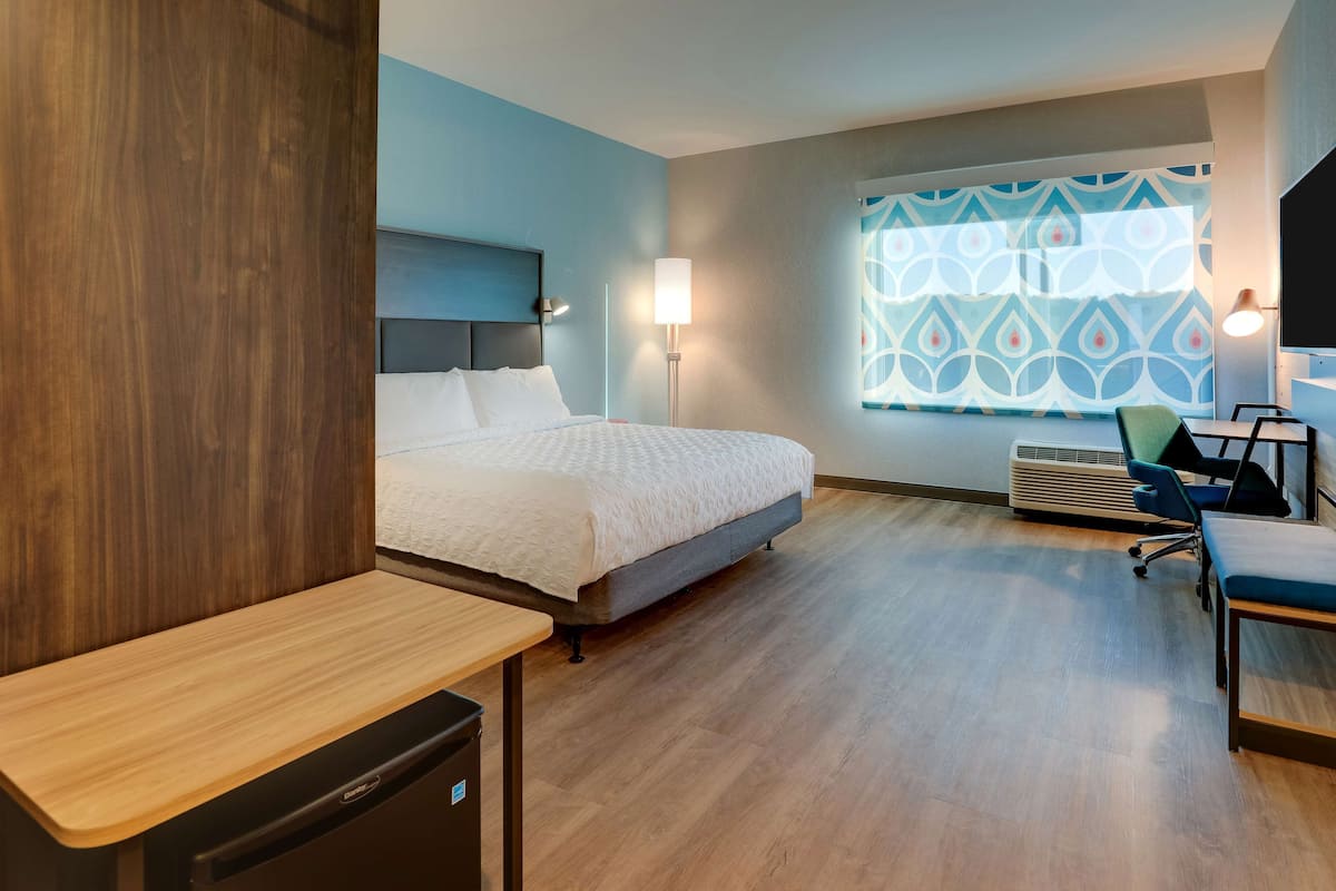 A hotel room with a bed, bedside table, desk, chair, lamp, air conditioning unit, and a window with a geometric pattern.