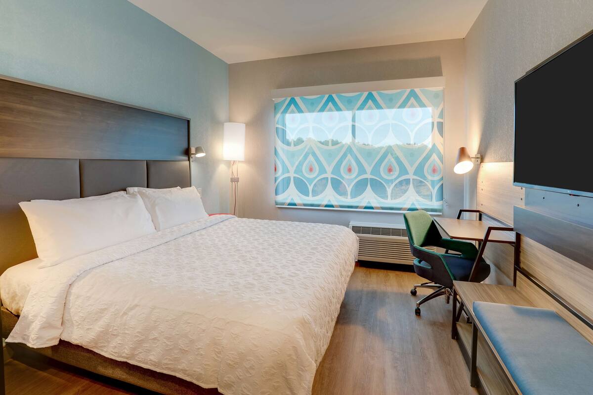 A hotel room with a large bed, a desk, a chair, and a large window with a geometric pattern.