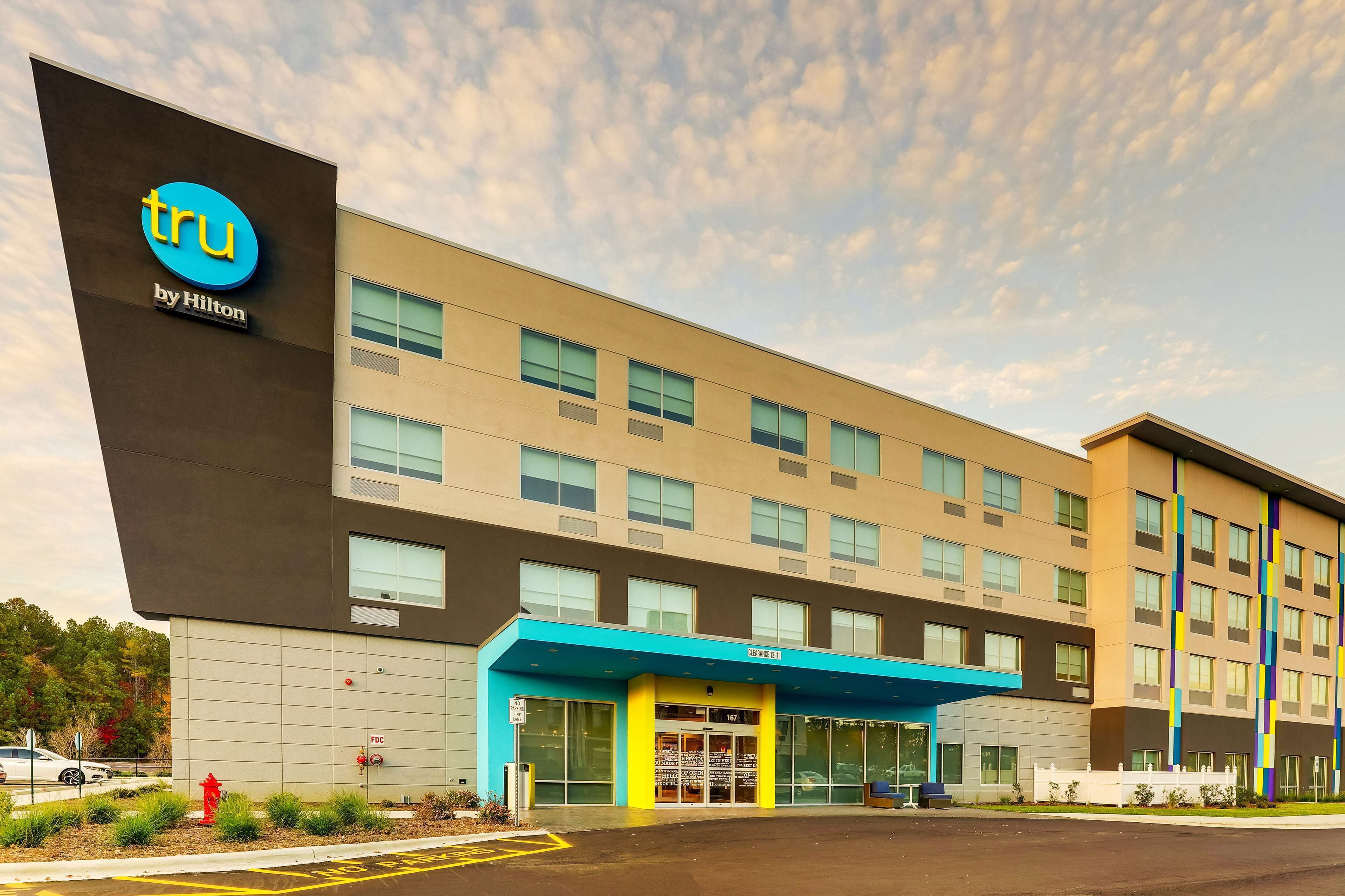 Un hotel de diseño moderno, con un llamativo letrero de 'tru by Hilton', un toldo de entrada azul y un estacionamiento.