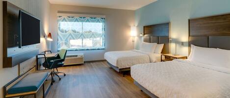 A hotel room with two beds, a desk with a chair, a TV, and a window with patterned curtains.