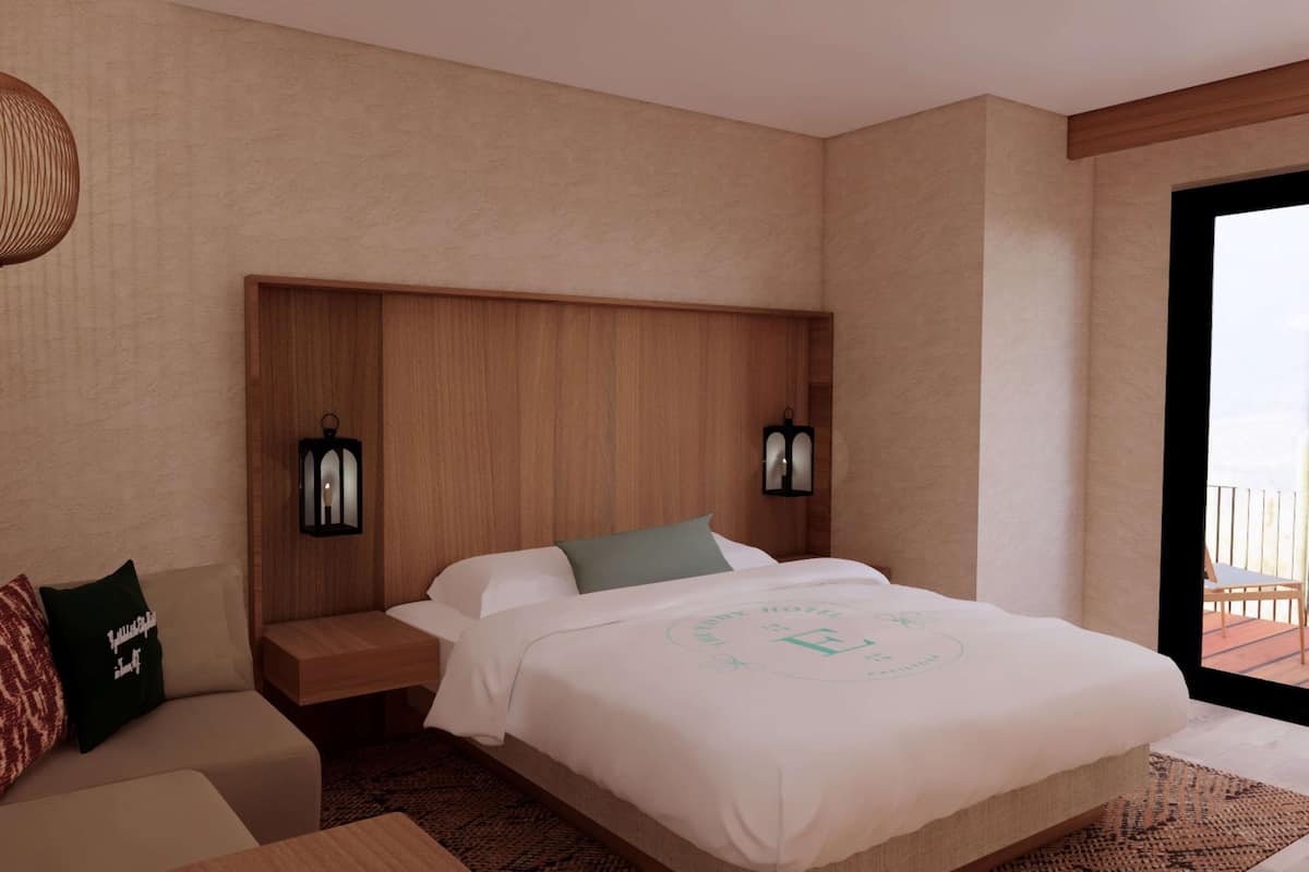 A modern hotel room with a large bed, a wooden headboard, two wall-mounted lamps, a sofa with patterned cushions, and a view of the outdoors through a glass door.
