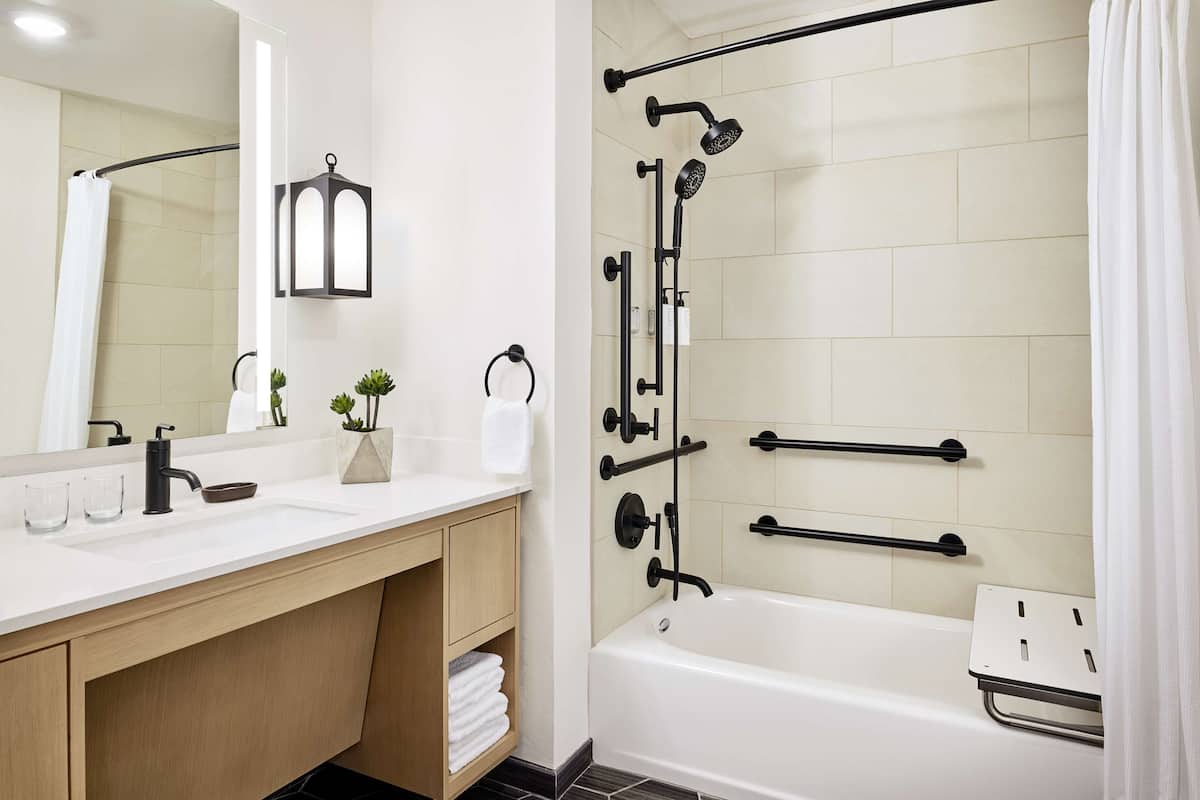 A modern bathroom with a white sink, mirror, and bathtub. There are two towel racks, a showerhead, and a small potted plant on the vanity.