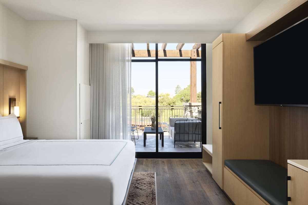 A hotel room with a bed, a TV, a balcony with a table and chairs, and a view of the outdoors.