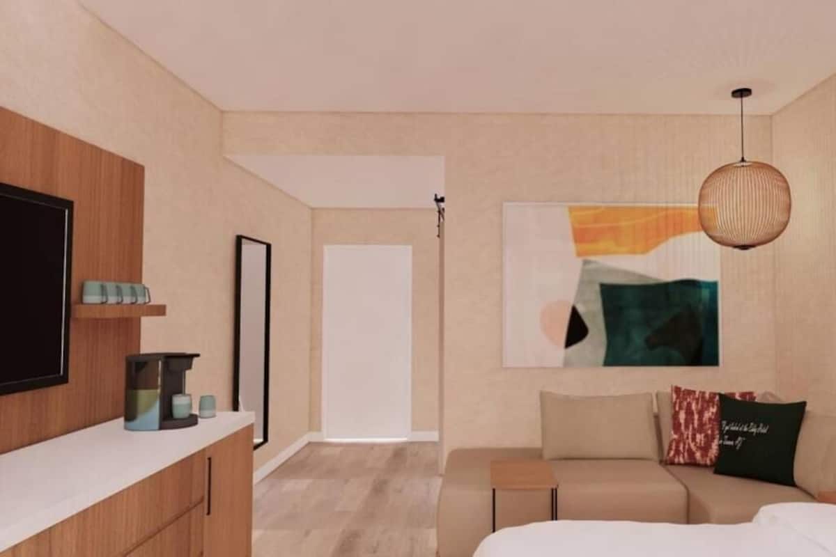 Moderni hotellihuone, jossa on sänky, sohva, televisio, kahvinkeitin ja taulu seinällä.