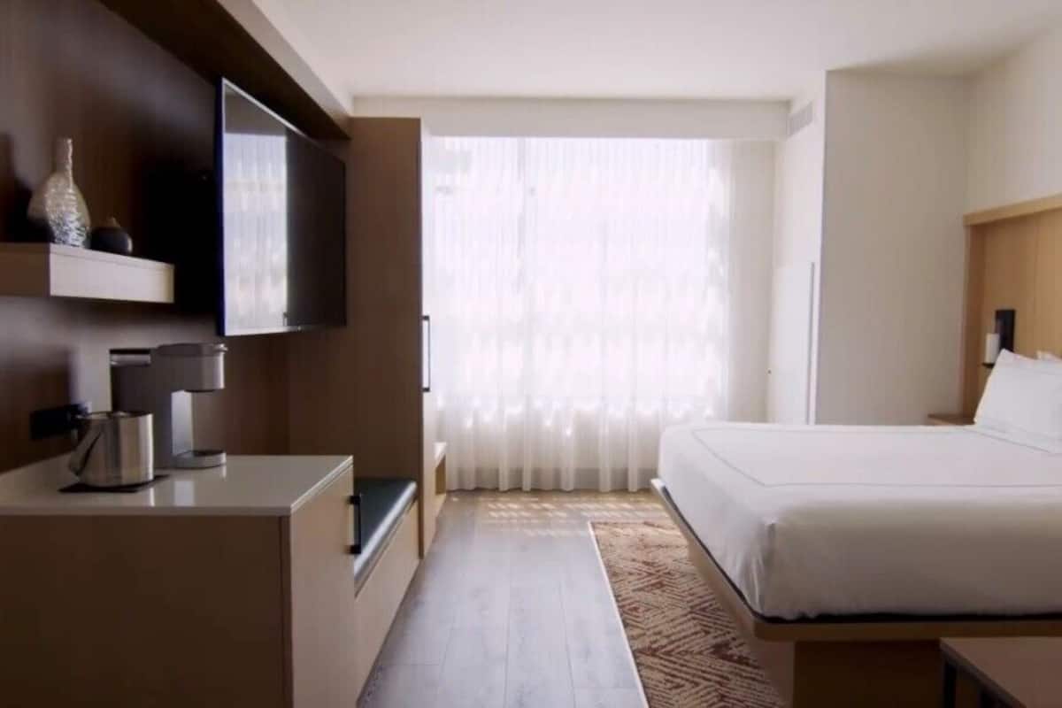 A hotel room with a bed, a TV mounted on the wall, a coffee machine, and a window with sheer curtains.