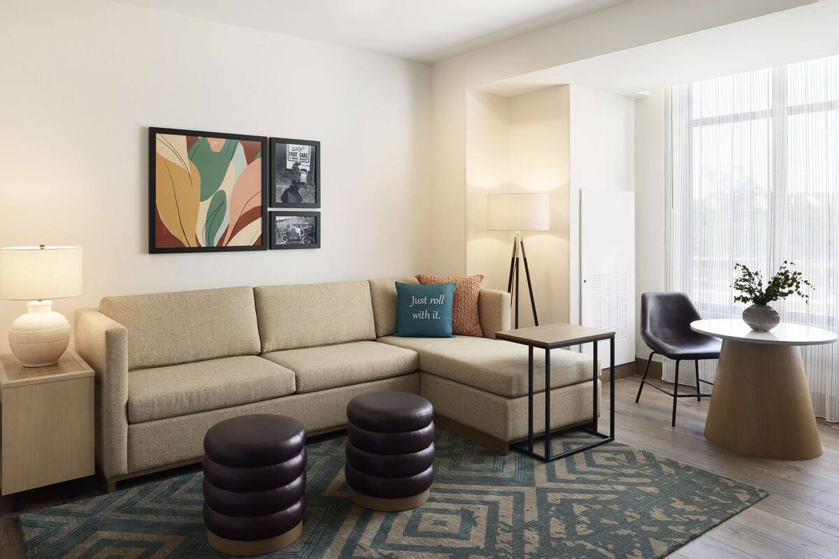 A modern living room with a sectional sofa, a coffee table, a side table, and a lamp. There are two ottomans, a rug, and a potted plant on the floor.