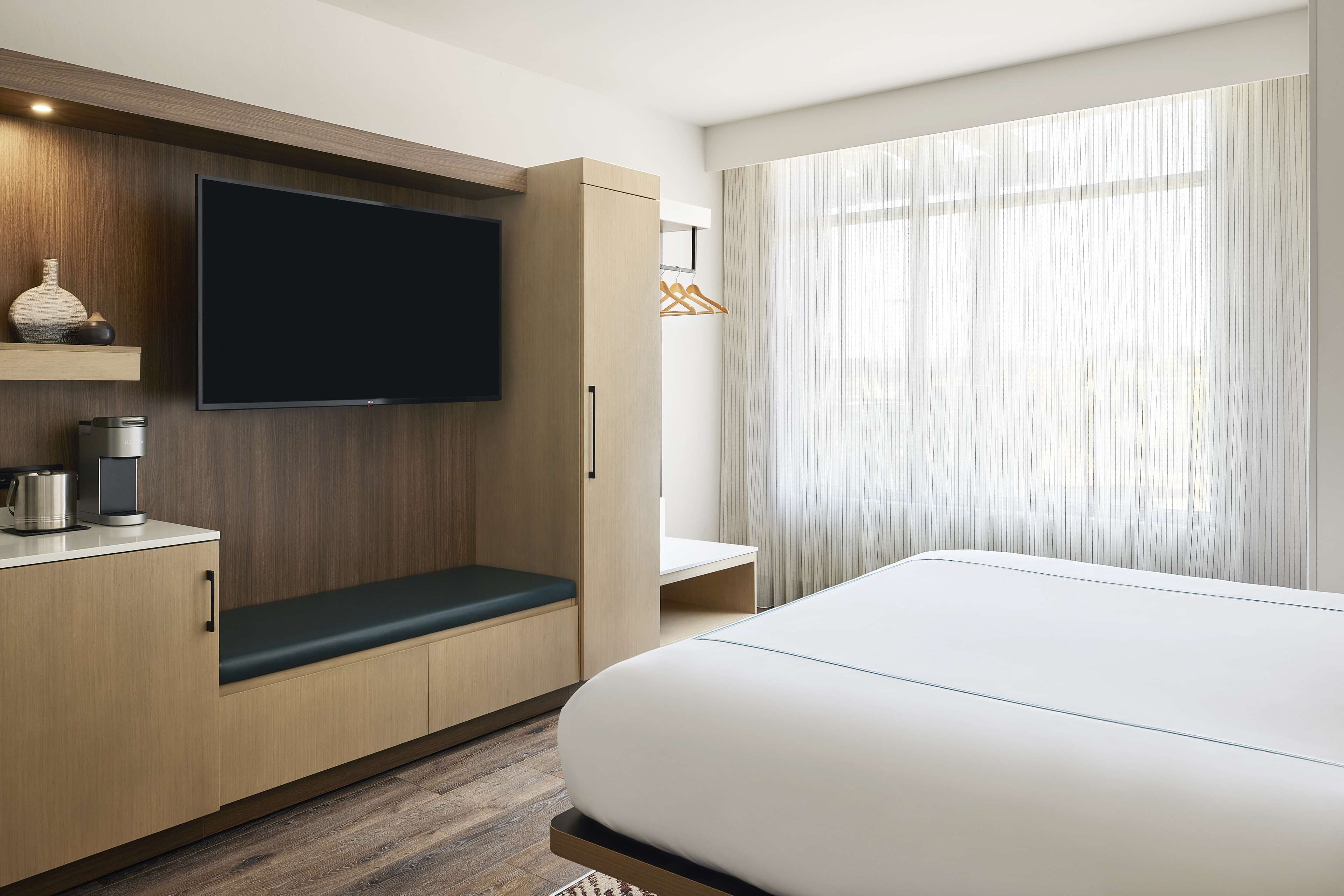 Une chambre d’hôtel moderne avec un grand lit, un téléviseur à écran plat fixé sur un panneau en bois, un banc avec un coussin vert, une petite table avec une machine à café et une grande fenêtre avec des rideaux légers.