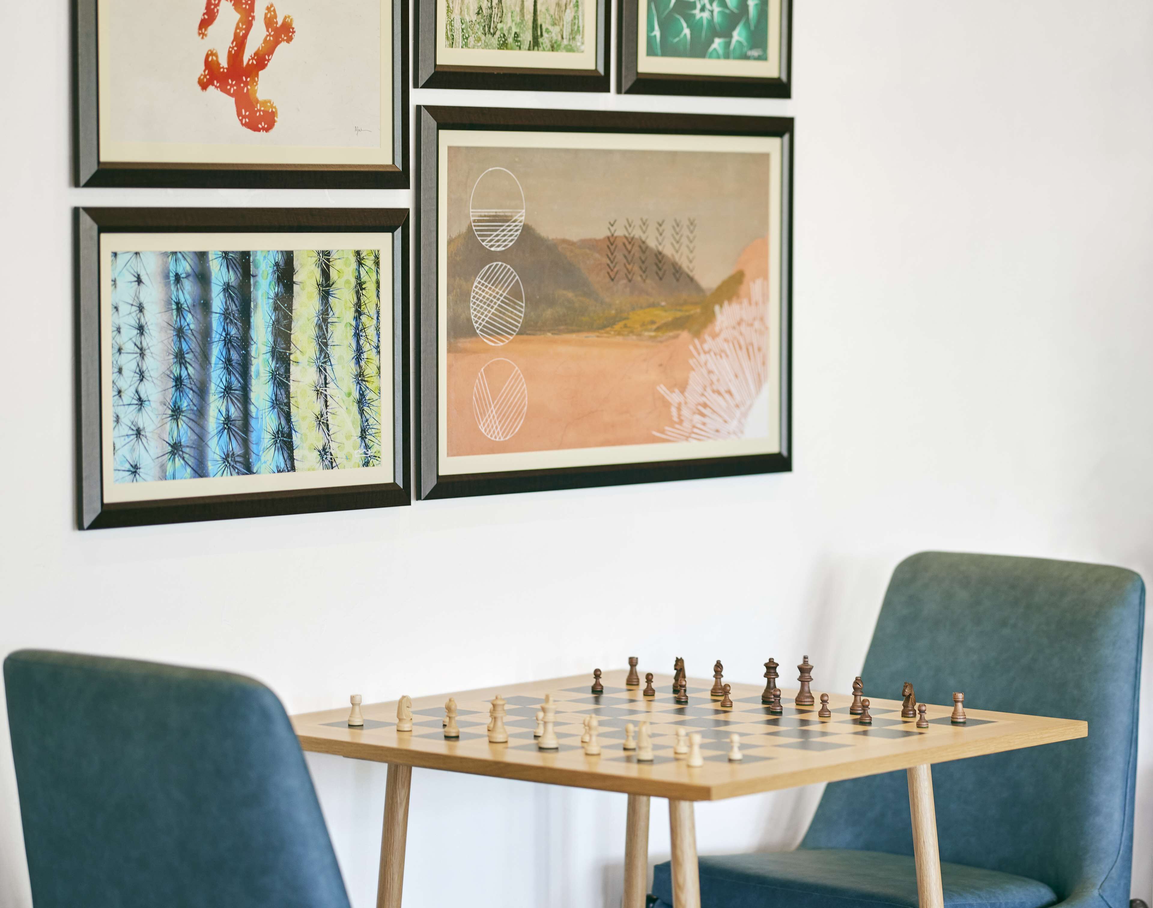 Un jeu d’échecs est posé sur une table en bois, avec deux chaises bleues, le tout étant entouré d’œuvres d’art encadrées accrochées au mur.