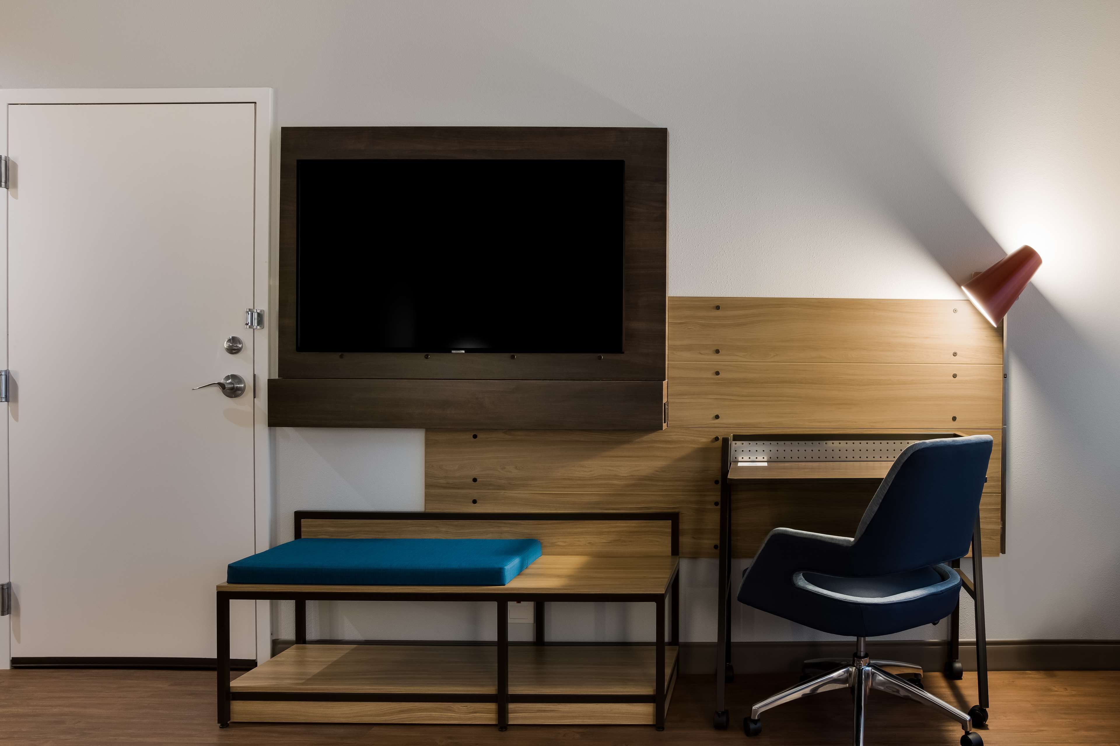 A room with a TV mounted on a wooden wall, a desk with a chair, and a bench.