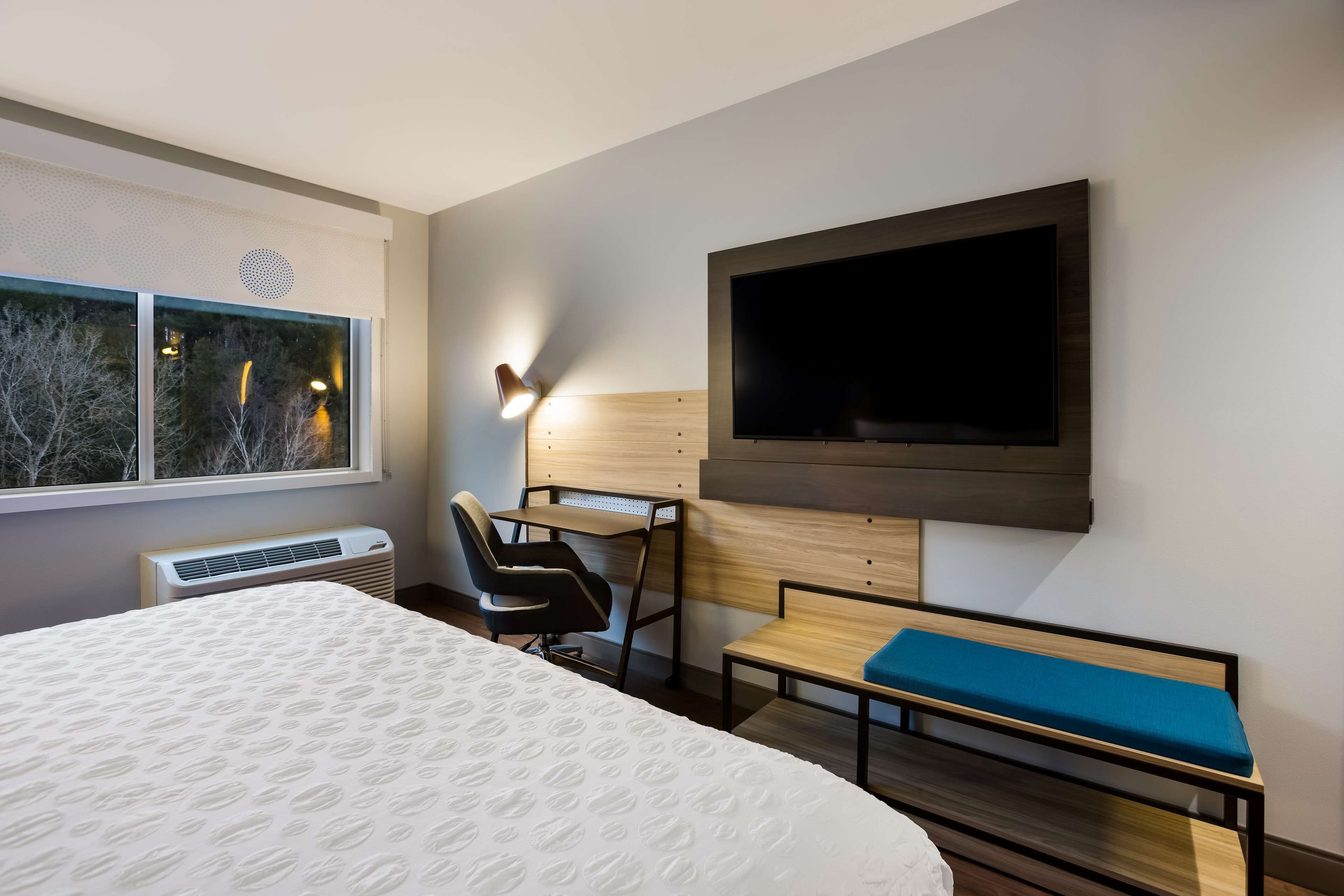 Ein modernes Hotelzimmer mit einem großen Bett, einem Schreibtisch mit Stuhl, einem an der Wand befestigten Fernseher und einem Fenster mit Blick auf Bäume.