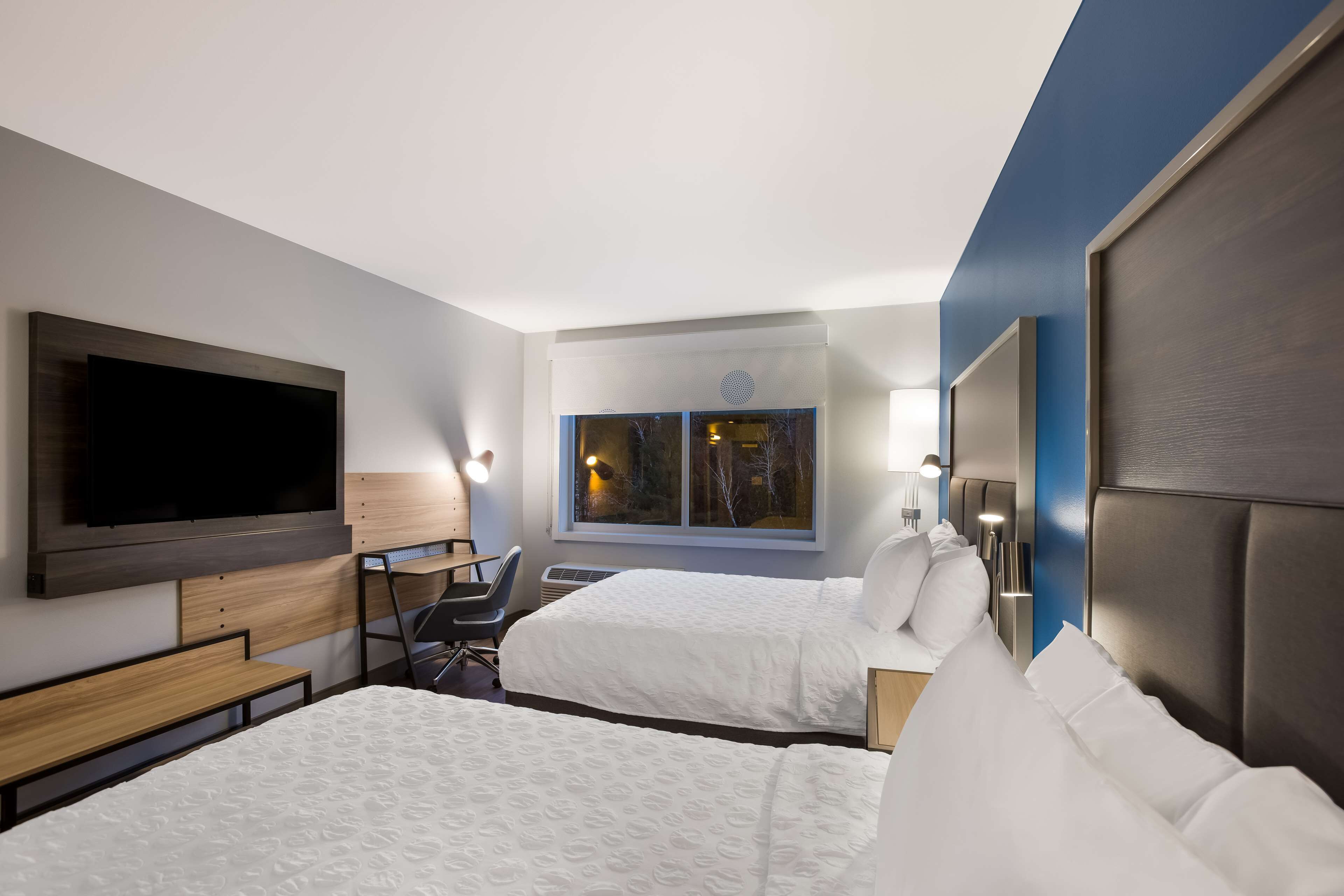 A hotel room with two beds, a TV, a desk, and a window with a view of trees.