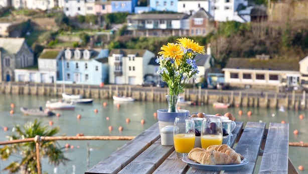 A wooden picnic table with a plate of croissants, glasses of orange juice, and a vase of flowers. - A bright and spacious three bedroom house with panoramic views over Looe. (Looe)