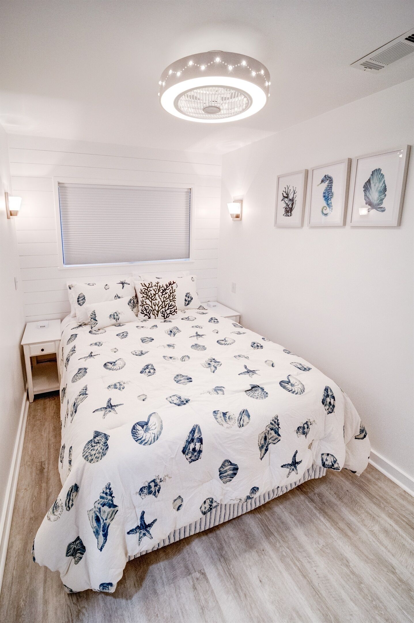A bedroom with a bed, a nightstand, a wall-mounted light fixture, and framed artwork on the wall.