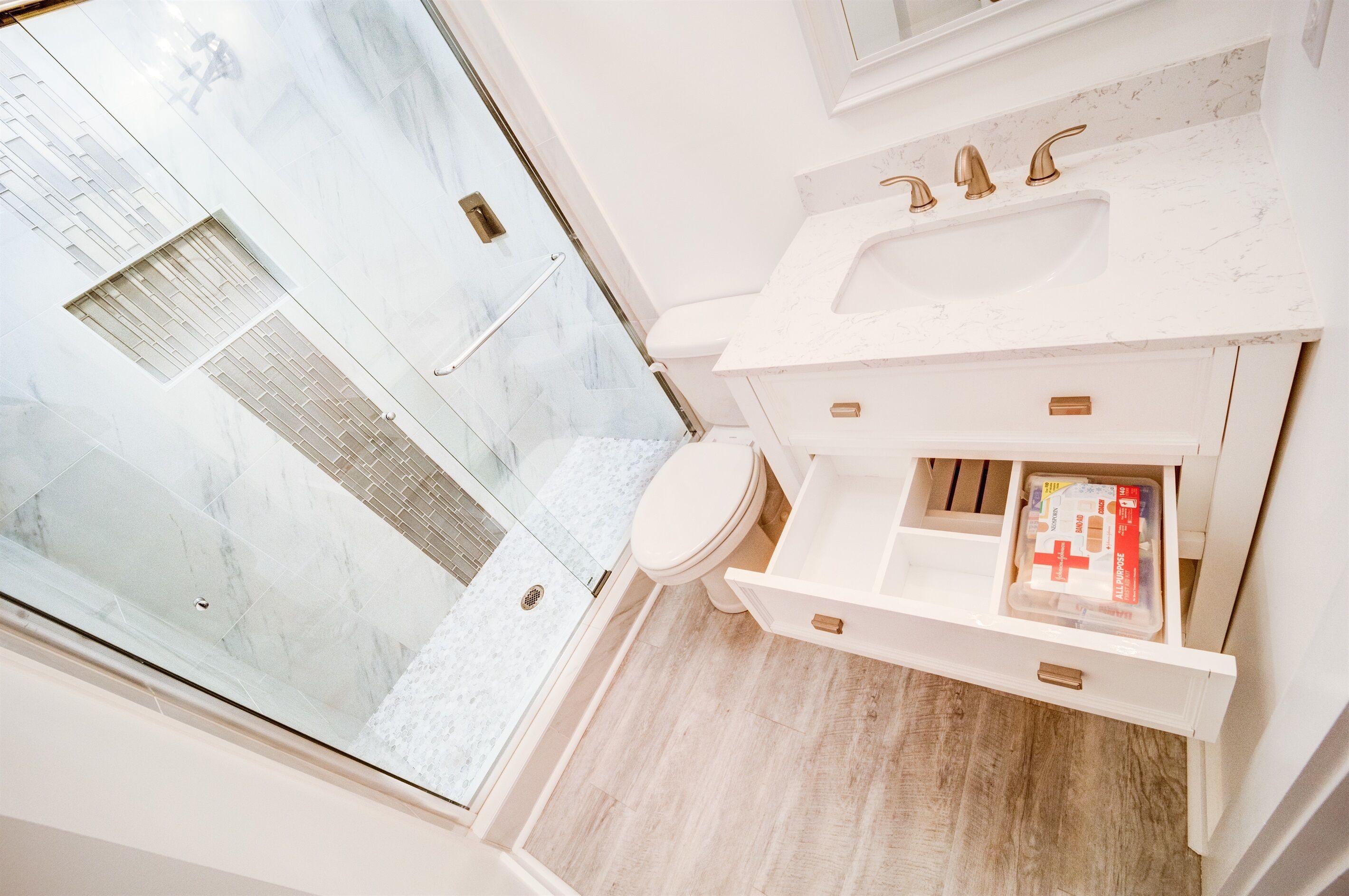 Un baño moderno con cabina de ducha de vidrio, lavamanos blanco con detalles dorados y mueble de tocador de madera con cajones.