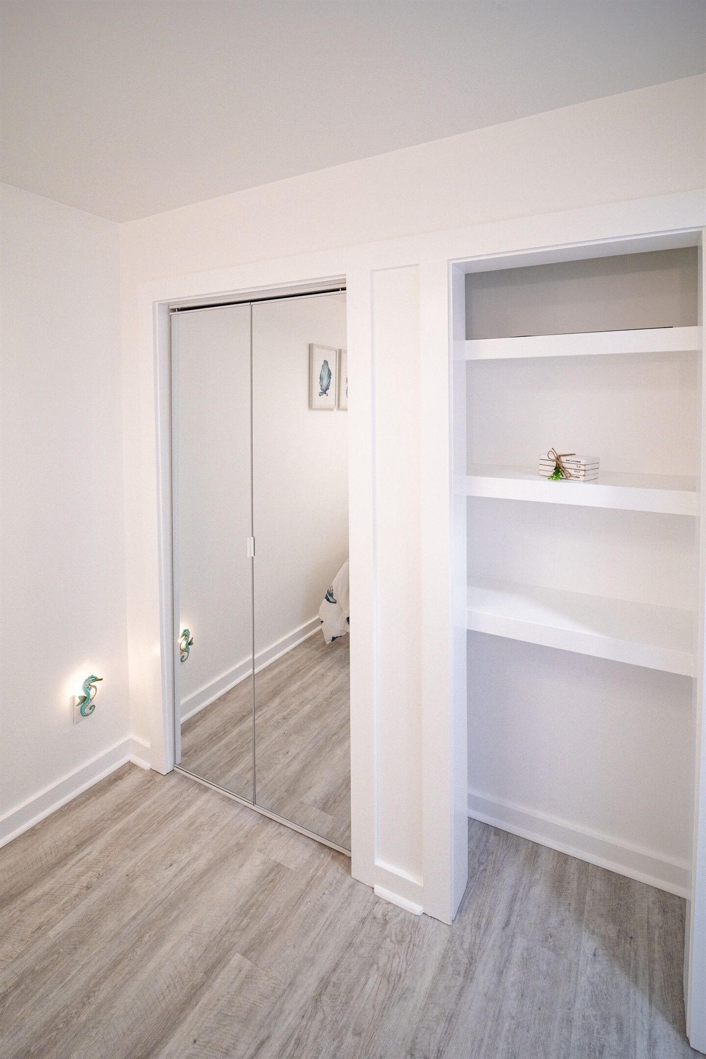 A room with a wooden floor, white walls, a mirrored closet door, and a white shelving unit with a few items on it.