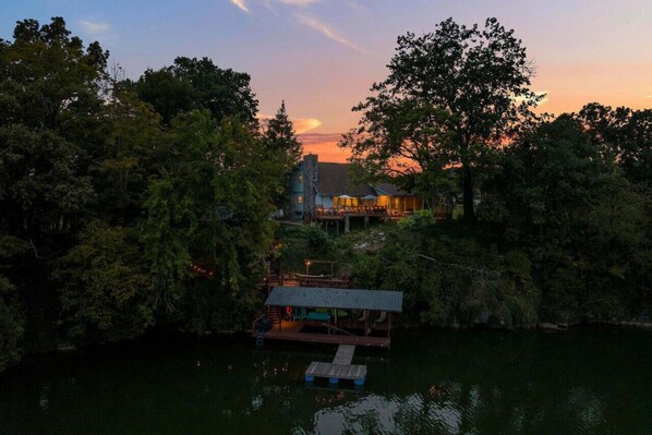 A lakeside cabin with a dock, surrounded by dense trees and a sunset in the background.