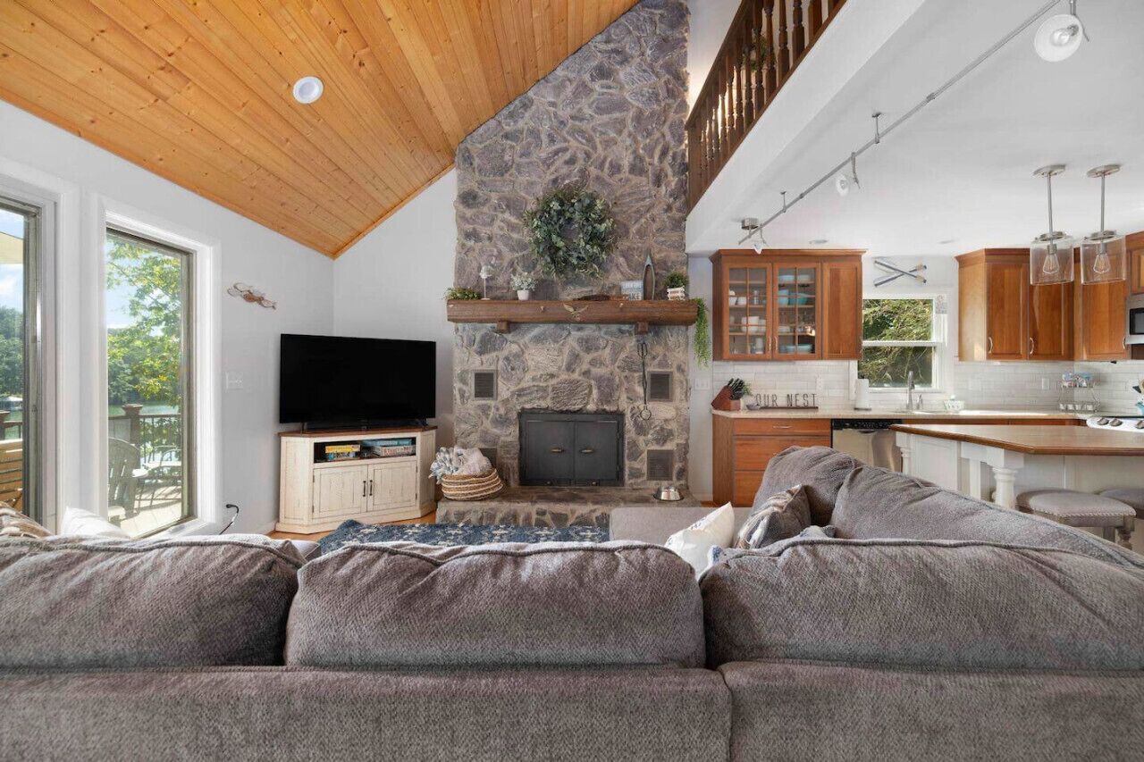 A modern living room with a stone fireplace, wooden ceiling, and a large window overlooking a deck.