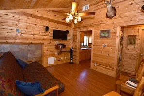 Interior de uma cabana de madeira com teto de madeira, um ventilador de teto, televisão montada, lareira de pedra, um sofá e uma área de jantar.