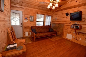 Interior de una cabaña de madera con un sofá, televisión y ventilador de techo.
