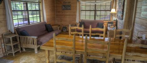 A cabin interior with a wooden dining table, chairs, a sofa, and a window with blinds.
