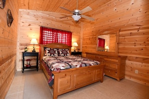 A wooden cabin bedroom with a bed, nightstands, a dresser, a ceiling fan, and a mirror.