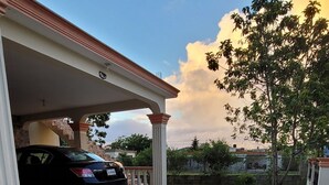 Un espacio al aire libre cubierto, con piso de piedra, un coche estacionado bajo una estructura blanca y un árbol al fondo.