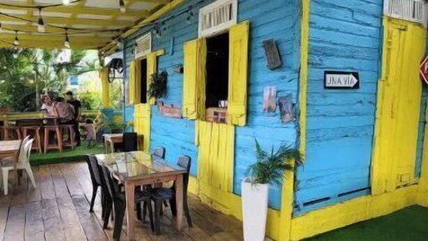 Un área de estar al aire libre con mesas y sillas de madera, un porche cubierto con vigas amarillas y un edificio azul con contraventanas amarillas.