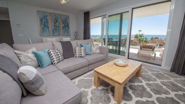 A modern living room with a grey sofa, patterned cushions, a wooden coffee table, and a large glass door leading to a balcony with a view of the sea. - Celere Dona Julia 2.3.0b (Casares)