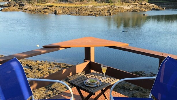 Une terrasse en bois avec deux chaises bleues et une table, donnant sur un lac paisible, avec une forêt en arrière-plan.