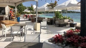 An outdoor seating area with white umbrellas, tables, and chairs, overlooking a beach and turquoise waters.