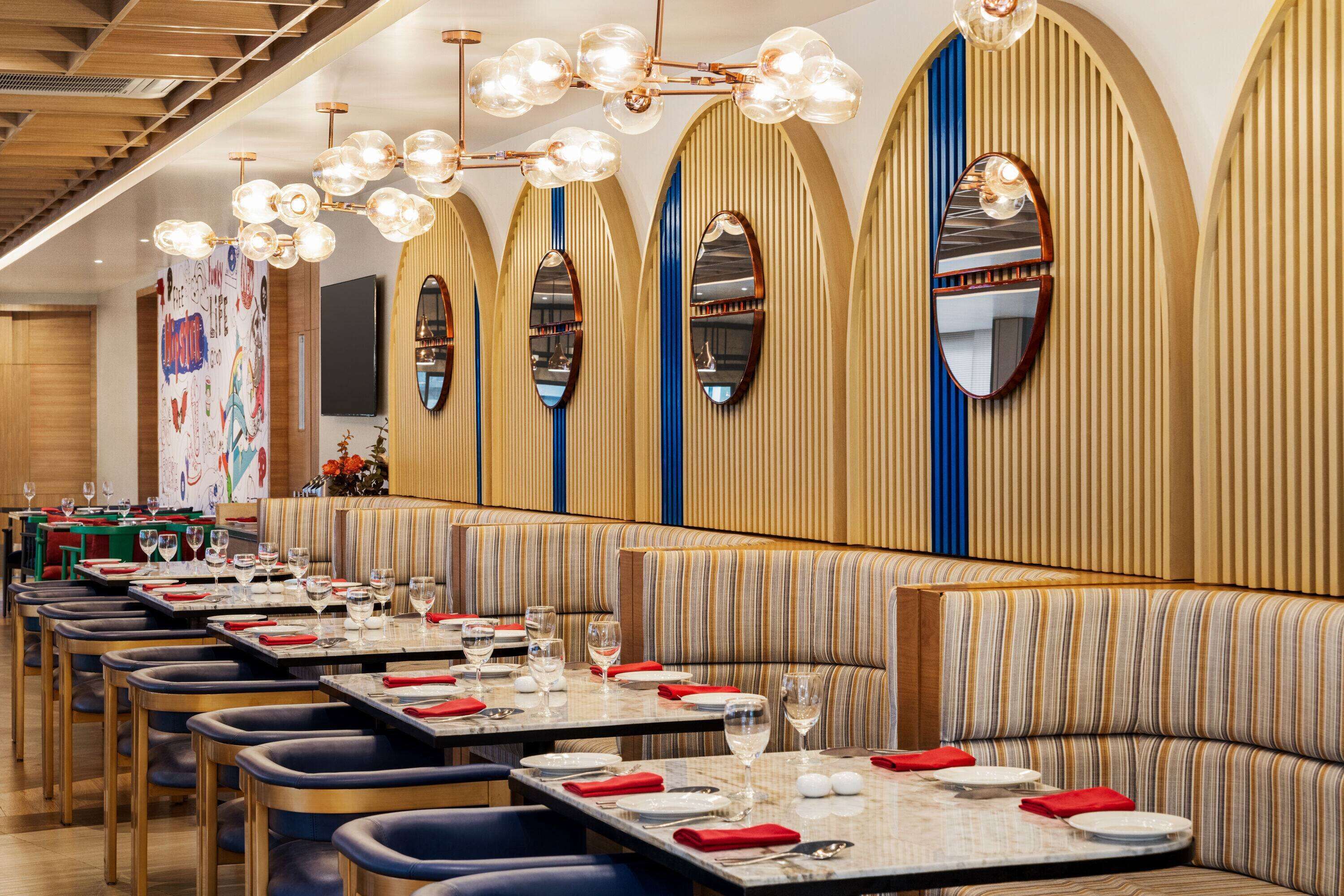 Un restaurant avec des banquettes à rayures, des tables dressées avec des serviettes rouges et des verres à vin, et un lustre moderne.