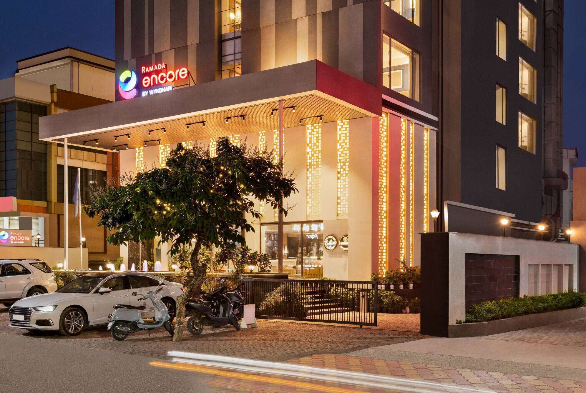 Una entrada moderna de hotel con un área exterior cubierta, iluminada con luces colgantes, y un letrero de "Ramada Encore by Wyndham".