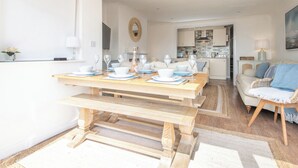 A dining area with a wooden table set for four, a sofa, and a kitchen in the background. - Coast View Apartment (Lyme Regis)