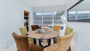 Un comedor con una mesa redonda de vidrio rodeada por seis sillas, una lámpara de techo y ventanas amplias con persianas.