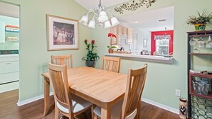 A dining area with a wooden table and chairs, a hanging light fixture, a framed photograph on the wall, and a kitchen in the background. - Close to Pelican Heron Golf Course and only  7 minutes to Lake Sumter Landing (The Villages)