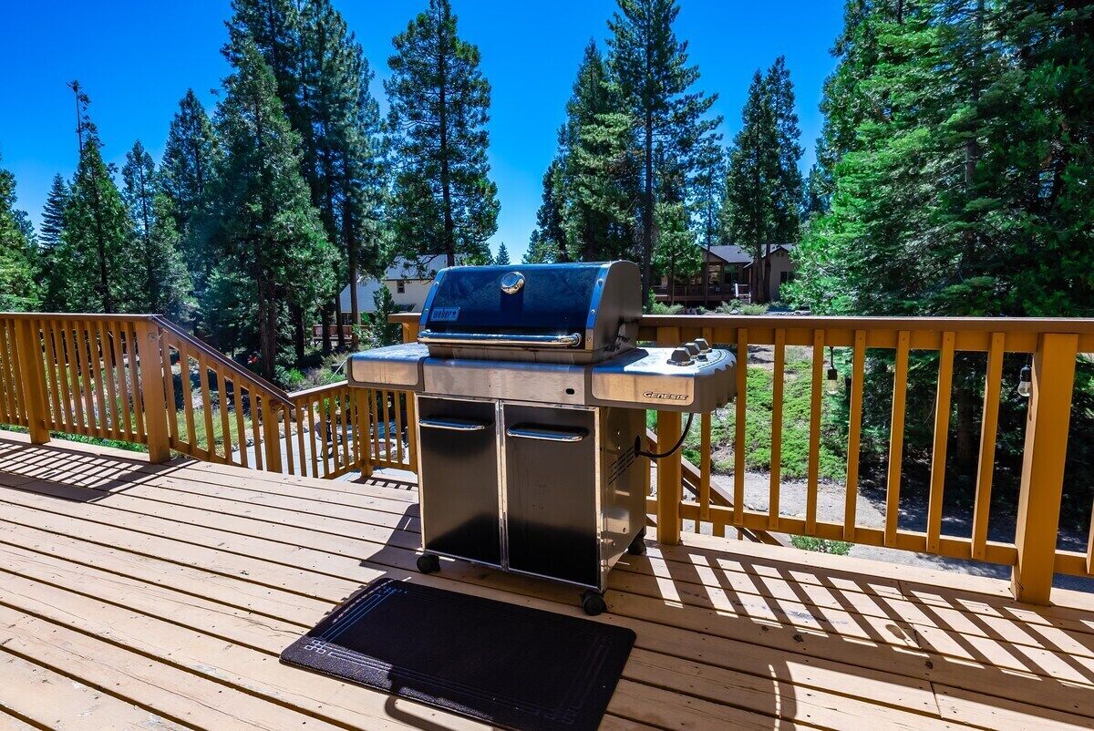 A barbecue grill on a wooden deck with railings, surrounded by tall trees.