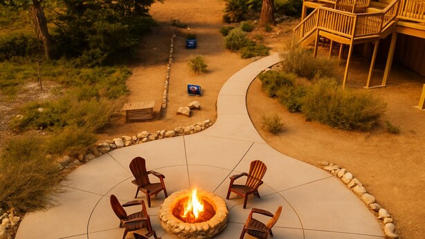 A fire pit surrounded by chairs on a patio with a stone border, a wooden deck, and a pathway leading to a building. - The Restful Rose ~ Fireside Vibes + AC Comfort (Shaver Lake)