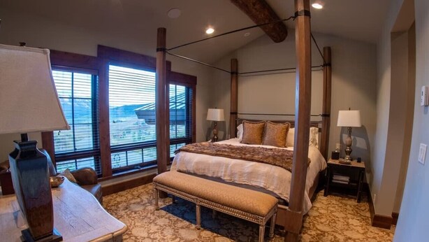 Un dormitorio con una cama grande, dos mesitas de noche, un banco y una ventana con persianas.