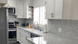 A modern kitchen with white cabinets, a stainless steel refrigerator, a microwave, and a stove. There is a window with blinds, a sink, and a countertop with a soap dispenser. - 3 Properties From the Beach (Newport Beach)