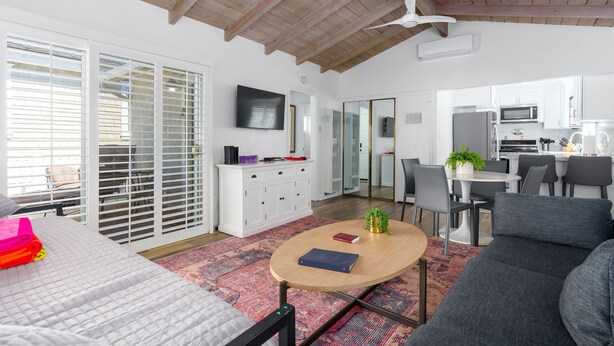 A modern living room with a sofa, a coffee table, and a TV. There is a kitchen area with a microwave and cabinets. - 3 Properties From the Beach (Newport Beach)