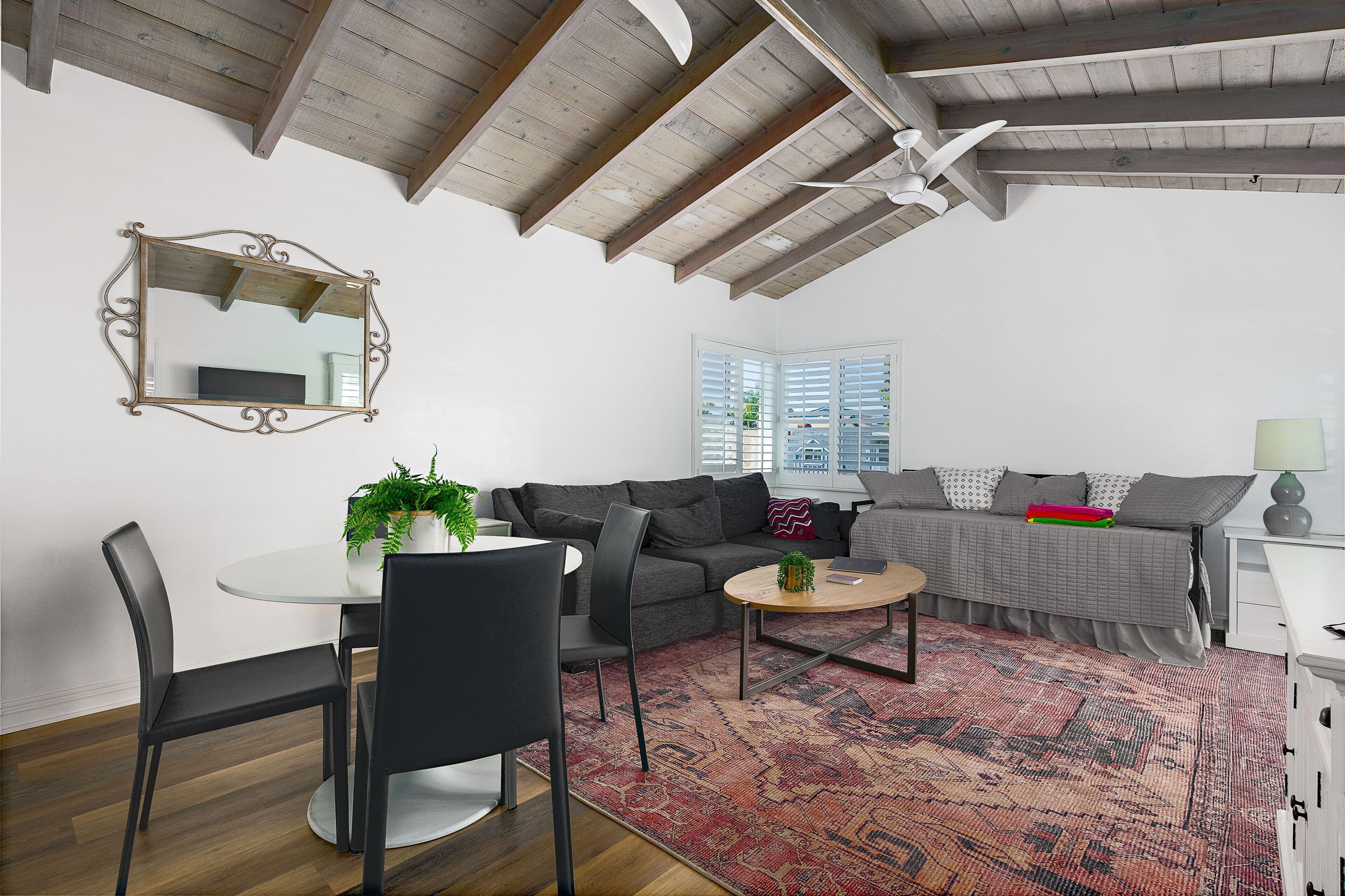 A modern living room with a dining area, a ceiling fan, a large mirror, and a view of the outside through a window.